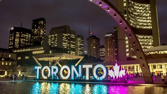 People enjoying the evening in front of and around the large outdoor Toronto sign , which is lit with multicolours as the center piece for the city hall, surrounded with tall buildings.